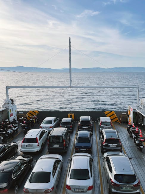 A view from inside a vehicle ferry showing the car deck with multiple parked cars arranged in rows, including sedans and SUVs, secured with wheel chocks and safety barriers in black and yellow stripes. The vehicle deck is open-air with a metal railing at the edge, behind which is a body of water with gentle waves and distant land visible on the horizon under a partly cloudy sky. In the background, a tall metal mast with support wires is visible, indicating a ferry or transport vessel used for home relocation and furniture transport services. This scene is associated with travel routes to Eastham Ferry and Village, EASTHAM, supported by Man with Van Eastham’s removal and moving services, which facilitate packing, loading, and transport logistics for local house removals.