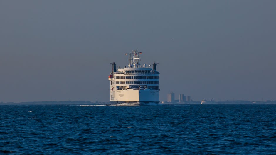 A large white passenger ferry approaching a calm body of water near the coast, with a distant city skyline featuring high-rise buildings visible in the background under a clear sky. The ferry’s front-facing design includes multiple decks and navigation equipment on the top. The surrounding water is deep blue with gentle waves, and the scene suggests a transportation route used for home relocation or moving services connecting Eastham Ferry and village, as managed by Man with Van Eastham, specialists in removals and furniture transport. The image captures the vessel in the process of arriving at or departing from the port area, illustrating the logistical aspect of house removals involving ferry routes, loading procedures, and the moving environment.