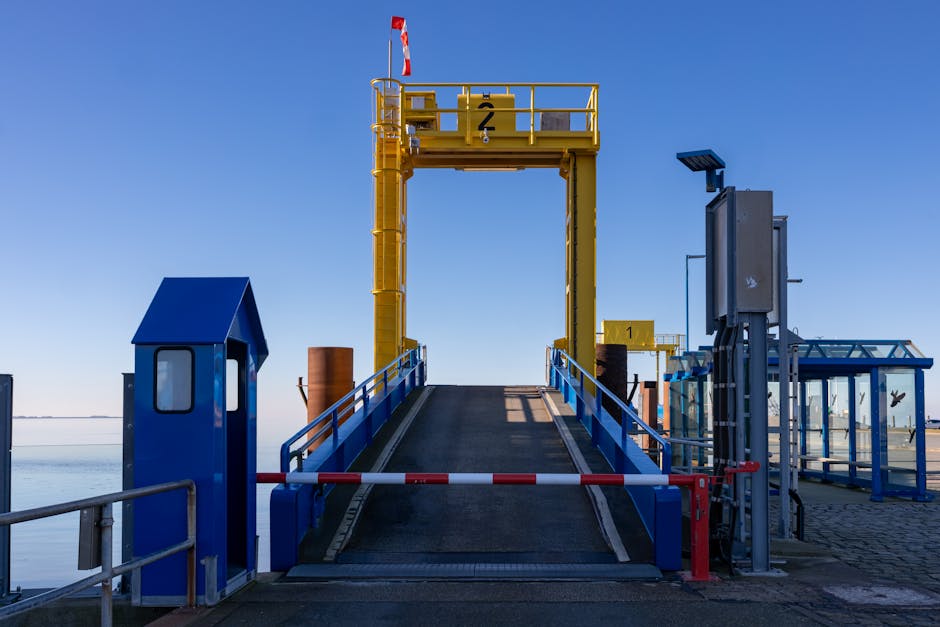 A view from inside a vehicle ferry showing the car deck with multiple parked cars arranged in rows, including sedans and SUVs, secured with wheel chocks and safety barriers in black and yellow stripes. The vehicle deck is open-air with a metal railing at the edge, behind which is a body of water with gentle waves and distant land visible on the horizon under a partly cloudy sky. In the background, a tall metal mast with support wires is visible, indicating a ferry or transport vessel used for home relocation and furniture transport services. This scene is associated with travel routes to Eastham Ferry and Village, EASTHAM, supported by Man with Van Eastham’s removal and moving services, which facilitate packing, loading, and transport logistics for local house removals.