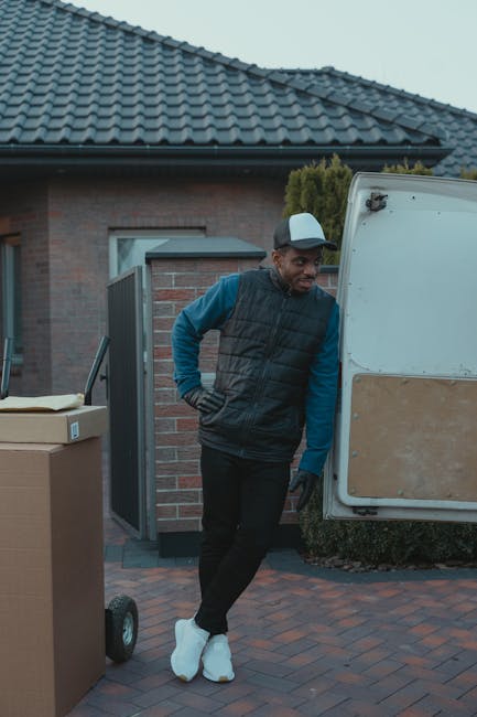 A man wearing a cap, black gloves, a black sleeveless jacket over a blue long-sleeved shirt, and black trousers stands beside a white moving van with a large wooden panel attached to its side. He is l