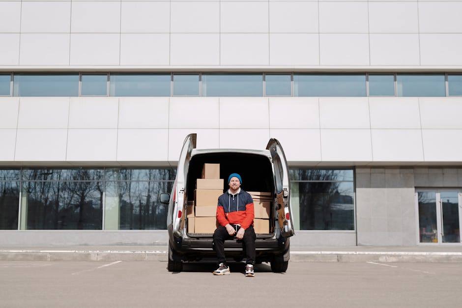 A man wearing a cap, black gloves, a black sleeveless jacket over a blue long-sleeved shirt, and black trousers stands beside a white moving van with a large wooden panel attached to its side. He is l