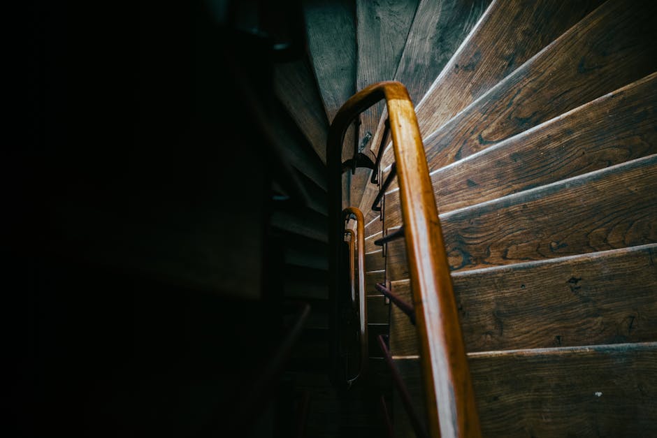 A wooden staircase situated inside a residential property, featuring dark brown, polished wooden steps and a matching wooden handrail. The staircase is narrow, with limited space, showing a steep ascent with clearly visible wood grain on each step. The photo is taken from the top of the stairs, looking down, capturing the vertical railing and the treads below. The environment is dimly lit, with natural light softly illuminating the wood surface, highlighting its texture and finish. The setting appears to be part of a home undergoing a house removal or furniture transport process. Occasionally, [COMPANY_NAME] handles such logistics, including navigating narrow staircases during packing and moving activities. The staircase is within an interior corridor, close to a dark wall on the upper part, emphasizing the confined space often encountered in house relocations, especially in terraced homes in Eastham.