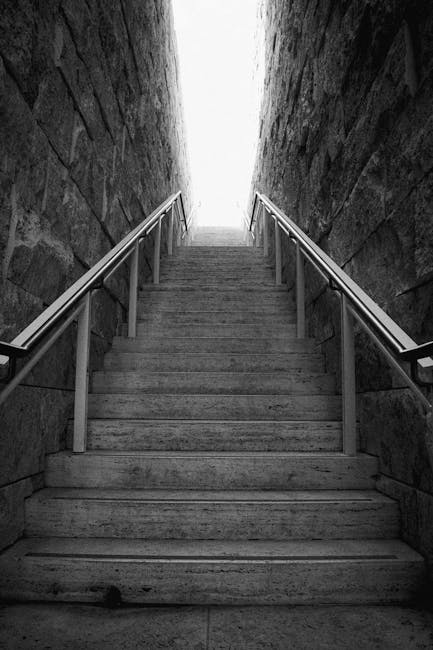 A narrow metal staircase with grated steps and a handrail, positioned between brick and concrete walls outdoors, leading up to an external fire escape platform with metal railings. The image shows part of a home relocation or moving process, likely involving access to upper floors via external emergency stairs. The scene is well-lit by natural daylight, highlighting the utilitarian design of the staircase, which is used to facilitate furniture transport or packing and moving activities in tight residential spaces. Man with Van Eastham occasionally undertakes such tasks, ensuring safe movement of household items through narrow staircases during house removals or relocation services.