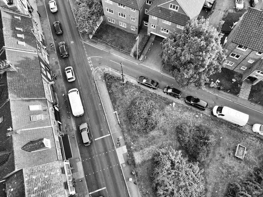 An aerial view showing a residential street with several parked cars lining the road and a white van, partially visible, parked on the pavement near a house with a front garden. The van is positioned close to the entrance of a property, with its rear doors open for loading or unloading furniture and boxes. Inside, there are cardboard boxes, wrapped furniture, and packing materials such as plastic wraps and fabric blankets, indicating a home relocation or moving process. Nearby, a trolley and moving straps are visible, used to assist with heavy items. The surrounding buildings are multi-storey houses with sloped roofs, and the street is well-lit with daylight. Man with Van Eastham’s vehicle is being prepared or used for furniture transport as part of a professional removals service, supporting efficient packing and loading for a household move on Wirral Council streets, Eastham known for adherence to permit rules for removal vans.