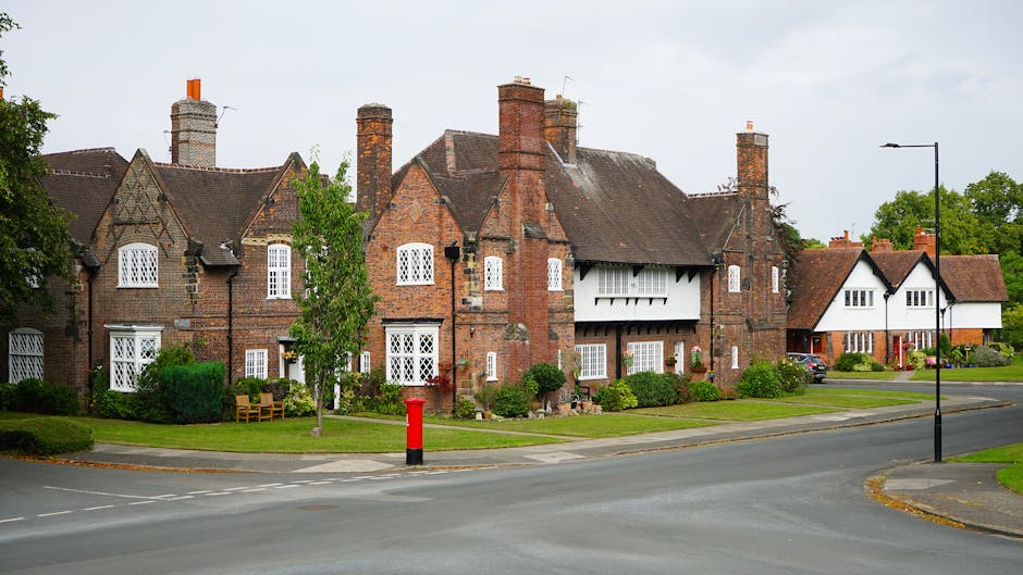 A row of traditional brick houses with pitched roofs and multiple chimneys, situated along a quiet residential street in Eastham. The houses feature white-framed windows with decorative glazing bars, and some have white sectioned or bay windows. The front gardens are well-maintained with trimmed bushes, small trees, and lawn areas; one garden includes a wooden bench. A narrow sidewalk runs parallel to the street, with a red post box positioned on the corner near the pavement. In the foreground, there is a black street lamp and a black traffic bollard with a red top. The scene is brightly lit with natural daylight, indicating a clear or partly cloudy day, and surrounding greenery suggests a peaceful suburban environment. This setting aligns with house removals or home relocation services provided by Man with Van Eastham, which are often involved in furniture transport and packing during local moves.