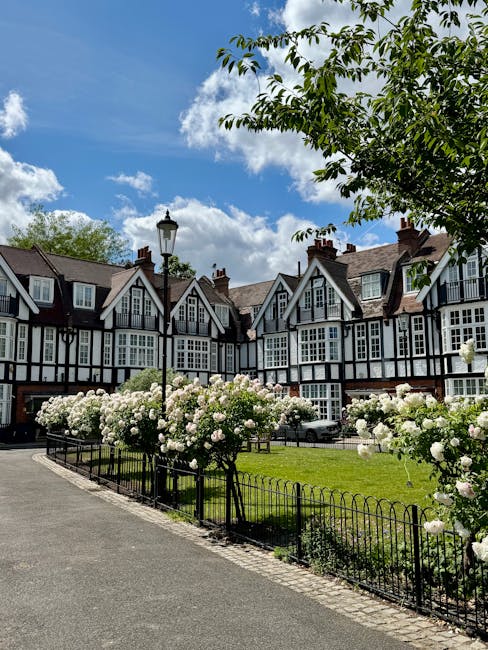 Photograph of a row of traditional multi-storey houses with black and white timber-framed facades, situated behind a small park area. The park features a paved pathway, a black metal fence, and a collection of blooming white roses along the fence. A tall, vintage-style street lamp stands near the pathway, and the scene is set under a bright blue sky with scattered clouds. The environment suggests a peaceful residential neighbourhood typical of the Port Sunlight estate, which may be part of a home relocation or moving process. Man with Van Eastham often handles furniture transport and packing services in such areas, ensuring careful loading and unloading inside the property or onto the van for a smooth house removal.
