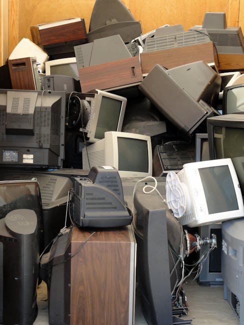 A large pile of old electronic items, including multiple vintage computer monitors with grey plastic casings, some with cords attached, stacked haphazardly inside a room with wooden walls. Among the monitors are old desktop towers, some with wooden veneer panels, and a few other electronic devices, such as a black telephone and a small radio. The equipment appears to be in a disorganized state, likely collected for disposal or recycling during a house clearance as part of a moving or home relocation process. The image captures the cluttered environment, suggesting the need for waste removal or collection of bulky items following a move. This scene exemplifies the type of waste management tasks carried out by companies like Man with Van Eastham during a home removal or disposal service, highlighting the importance of proper handling of household electrical waste during property clearance.