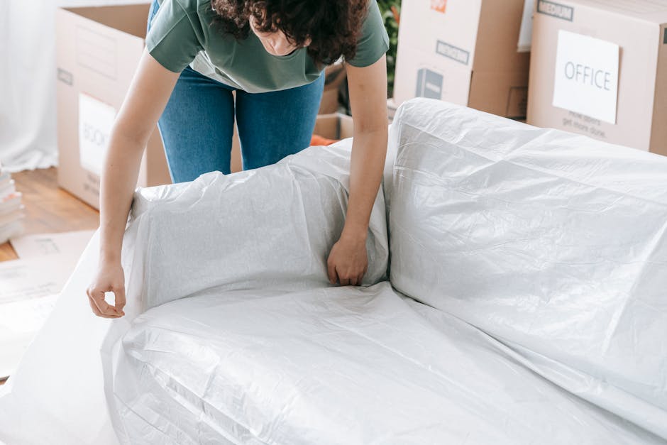 A person with curly hair wearing a green top and blue jeans is seen carefully wrapping a piece of furniture, likely a sofa or armchair, with white protective packing material in a room filled with moving boxes and cardboard crates. Some boxes are labeled with categories such as 'OFFICE,' indicating a home or office relocation process. The person is standing on a wooden floor, with a focus on the detailed task of preparing furniture for transport during packing and moving steps. In the background, there are additional packing supplies, including plastic wrap and cardboard boxes, arranged around the space, which appears to be part of a house or office being prepared for a home relocation or furniture transport. The scene reflects the careful handling and packaging involved in moving services offered by companies like Man with Van Eastham, highlighting the process of removing bulky furniture or waste after a move inside a residential or commercial property.
