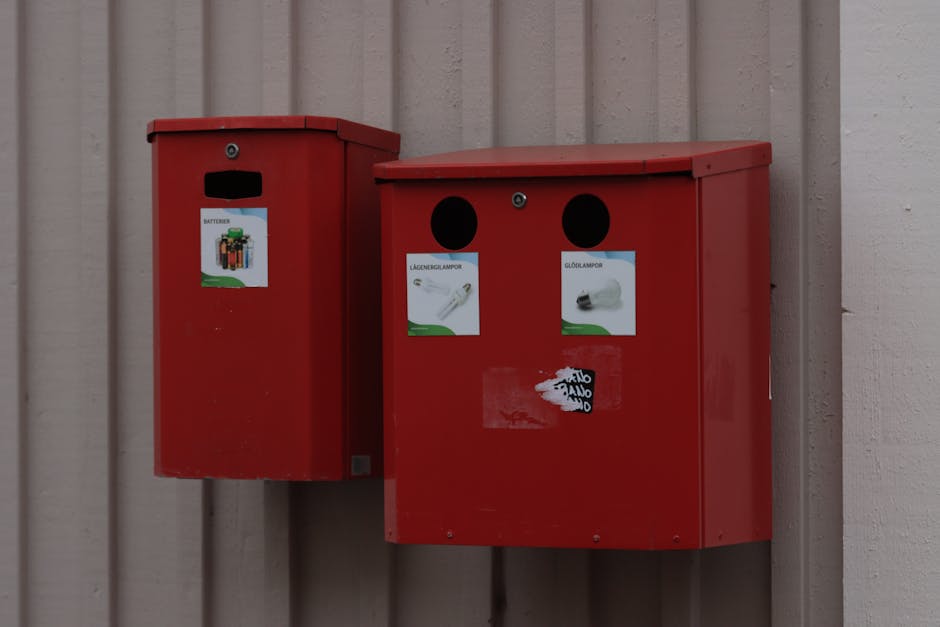 Two red communal or public waste collection bins mounted on an exterior beige wooden wall, with the larger bin on the right featuring two black circular openings and labels indicating recycling categories, including light bulbs and batteries, while the smaller bin on the left has a slit slot for general waste and a label with a battery symbol. The larger bin is positioned slightly to the right of the smaller one, both situated on a concrete or paved surface. The bins are part of a home relocation or packing process managed by Man with Van Eastham, supporting effective waste disposal and recycling laws in Eastham (Wirral). The lighting is natural and even, highlighting the clean, organized appearance of the bins and the wall behind them, essential for proper waste sorting during house removals or moving logistics.