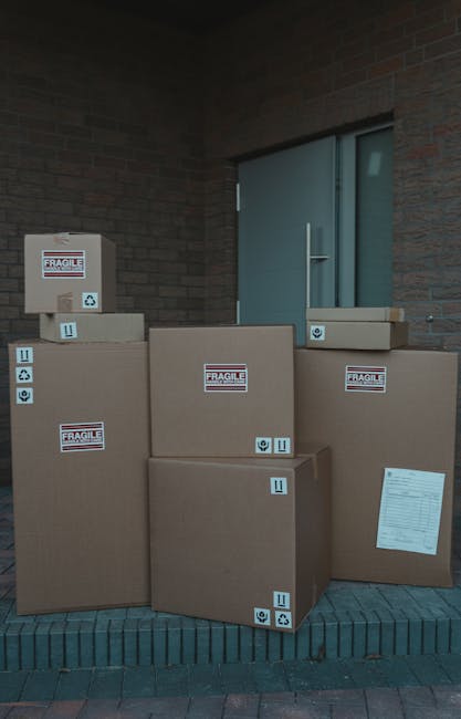 Several large cardboard boxes, some with 'FRAGILE' labels, are stacked outside a brick building near a blue door, positioned on a brick paved area. The boxes vary in size and are secured with packing tape, with some additional labels indicating careful handling. One of the boxes features a packing list attached. The setting suggests the packing stage of a home relocation or furniture transport process, as part of the services offered by Man with Van Eastham. The scene captures the preparation for a house move, where packed items are ready to be loaded onto a vehicle, potentially seen nearby or awaiting collection. The lighting is natural, and the arrangement emphasizes the organized approach to packing for an efficient removal or moving service.