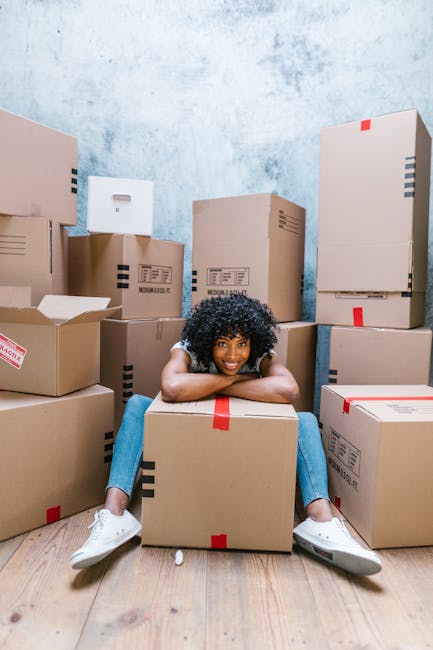 A young woman with curly black hair, wearing a white T-shirt, blue jeans, and white sneakers, is sitting on a light wooden floor surrounded by stacked cardboard boxes of various sizes. Some boxes are sealed with red tape, while others are open, revealing packing materials. She is smiling and resting her arms and chin on a large sealed box in front of her. The room has a textured light blue wall, and the scene suggests she is in the process of packing or preparing for a house move managed by Man with Van Eastham, with a focus on moving logistics, packing, and home relocation. The boxes are arranged around her, indicating an active packing or loading process as part of a house removal or furniture transport in the context of an urgent same-day move in CH62.